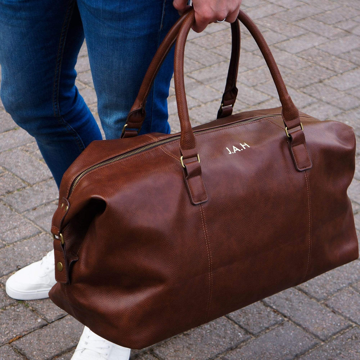 Man carrying a brown PU leather bag with personalised initials in gold by Original Monkey Gifts. Man wears blue denim jeans and white trainers.