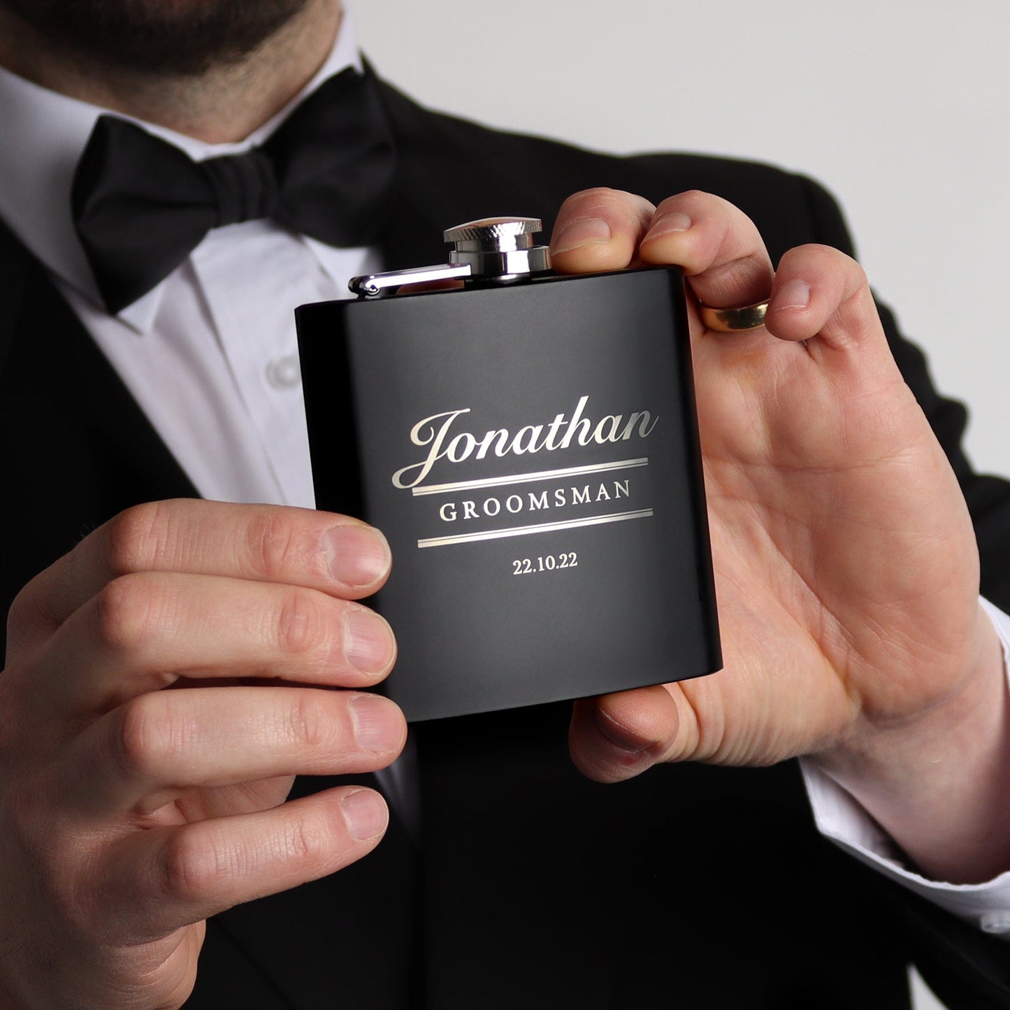 a man wearing a tuxedo hold a matt black hip flask with his name, role and wedding date engraved on the front, the perfect wedding gift for the groomsmen by handford and mason