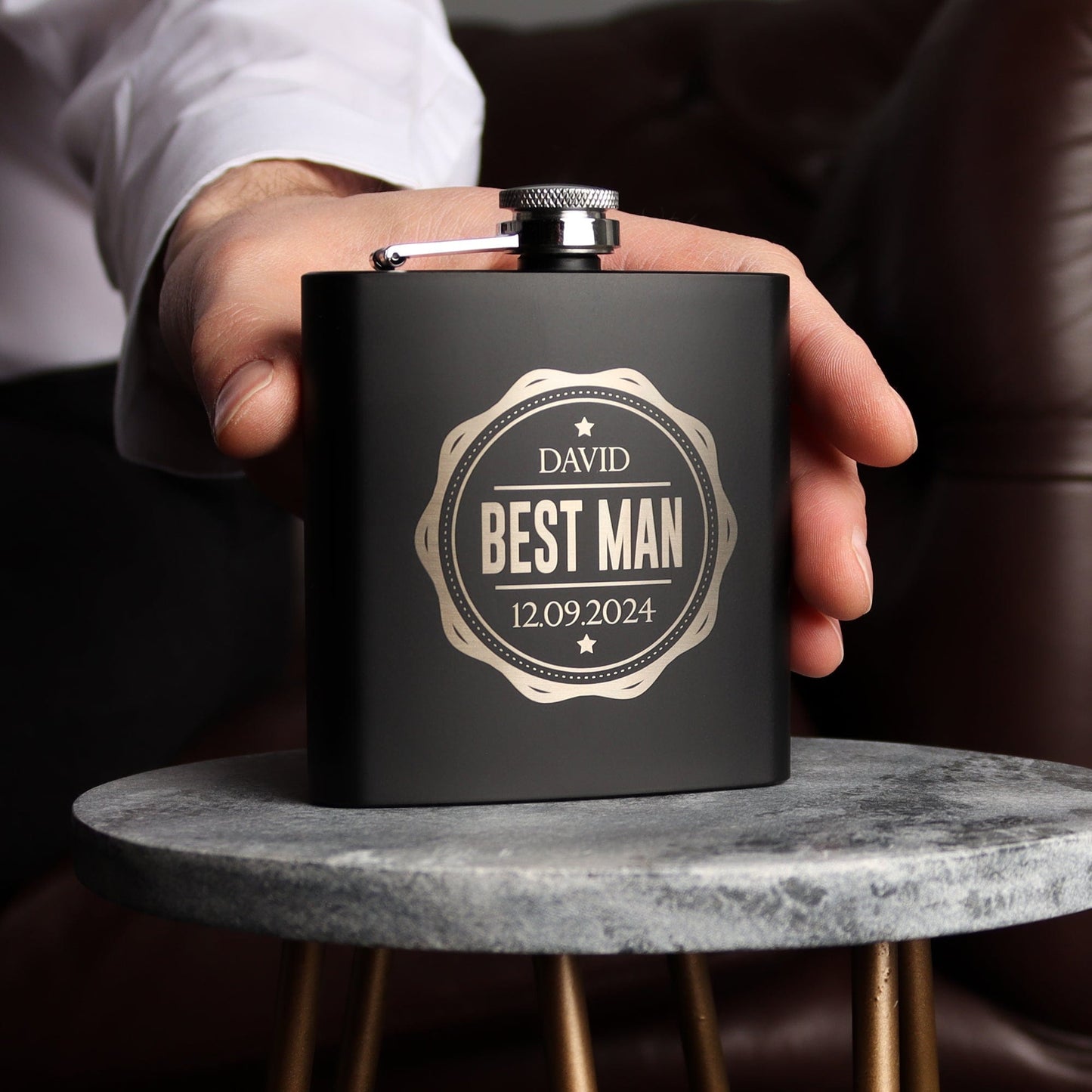 a man holding a black hip flask with engraving on the front in a stamp style, the engraving has his name, role and wedding date. Perfect groomsmen gift by handford and mason