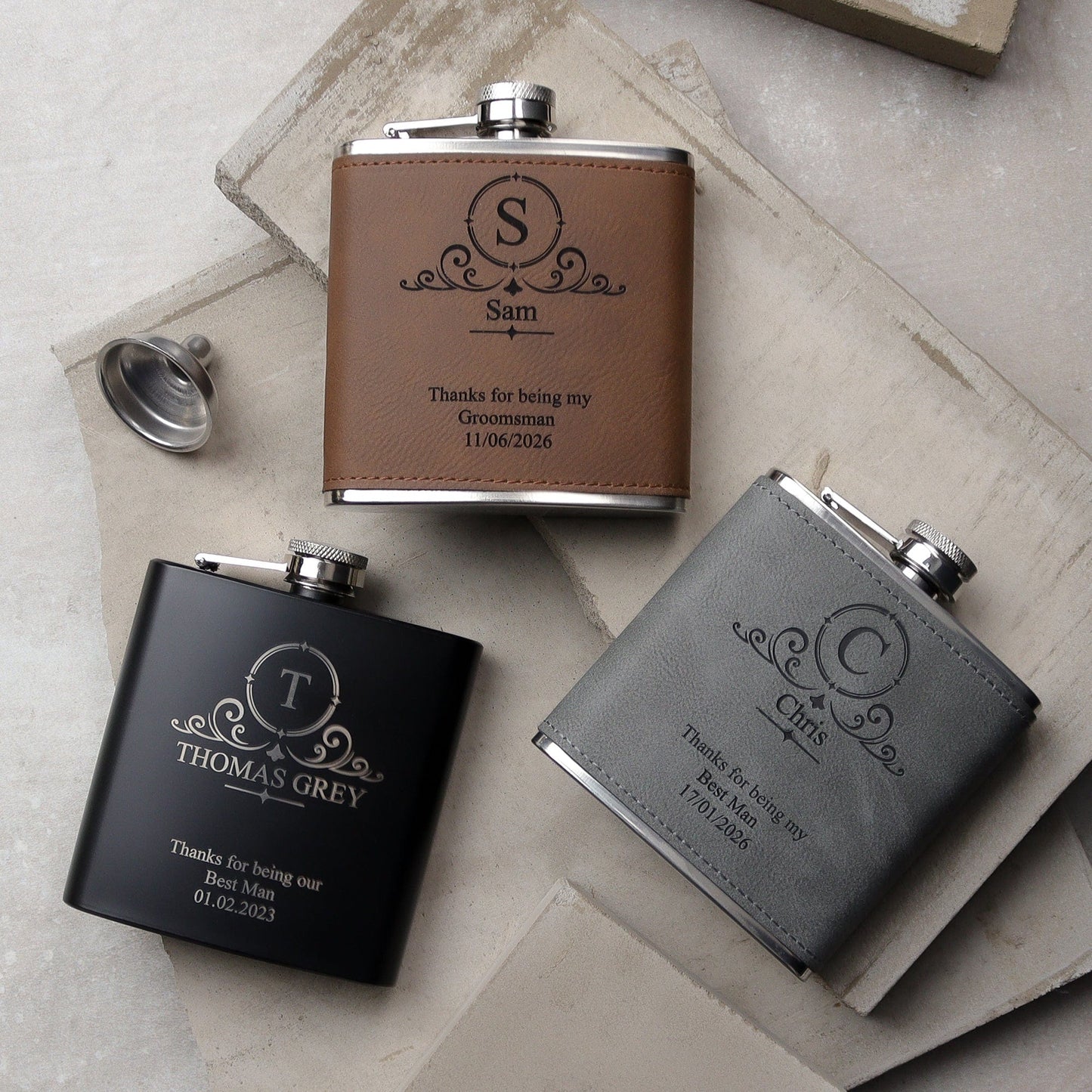 a set of three flasks all side by side one black, one grey, one brown all with a traditional design on the front with the recipients names, role and the wedding date all laser engraved. the perfect wedding groomsman gift.