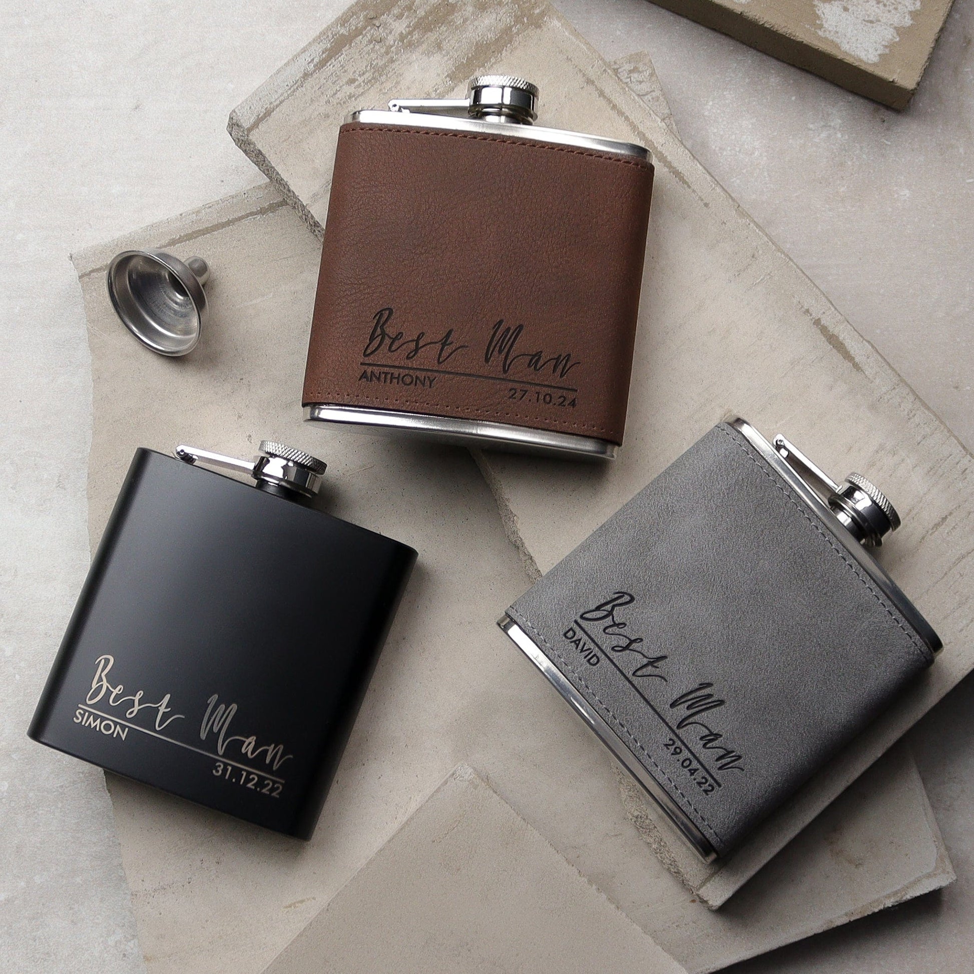 a set of three flasks all side by side one black, one grey, one brown all with a traditional design on the front with the recipients names, role and the wedding date all laser engraved. the perfect wedding groomsman gift.